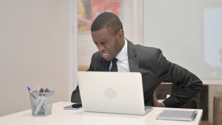 African Businessman having Back Pain while Using Laptop in Officeの写真素材