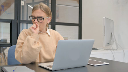 Coughing Female Worker Using Laptop in Officeの写真素材