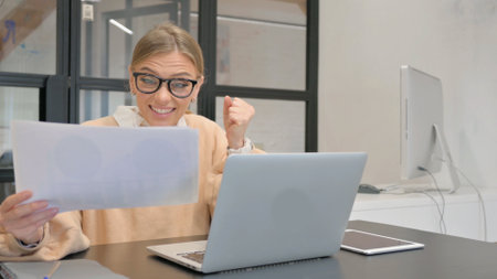 Female Worker Celebrating while Using Laptop and Documentsの写真素材