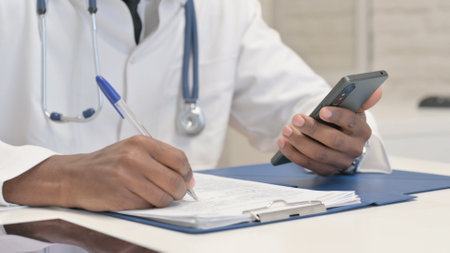 Close up of African Doctor Using Smartphone while Doing Paperwork in Hospitalの写真素材