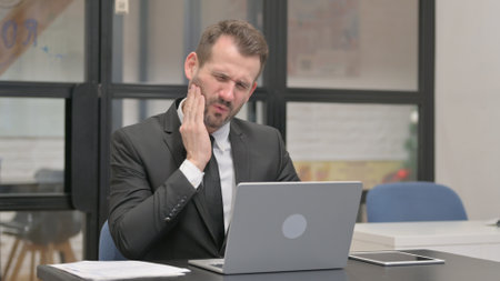 Businessman having Tooth Pain while Working on Laptopの写真素材