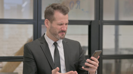 Portrait of Middle Aged Businessman Shocked by Loss on Phoneの写真素材