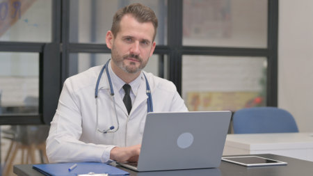 Middle Aged Doctor Looking at Camera while Working on Laptopの写真素材