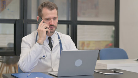 Middle Aged Doctor Talking on Phone and Using Laptopの写真素材