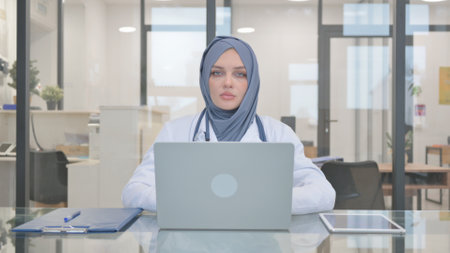 Doctor in Hijab Looking at Camera while Working on Laptopの写真素材