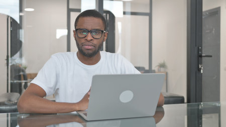 Young African Man Looking at Camera while Working on Laptop in Officeの写真素材
