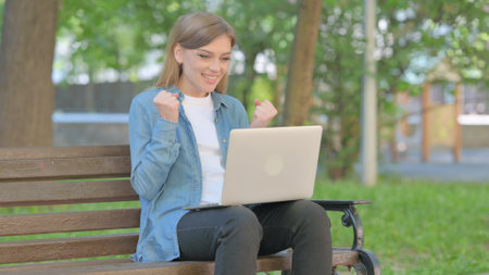 Outdoor Sitting Young Woman Celebrating Success on Laptopの写真素材