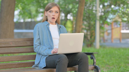 Shocked Young Woman Looking at Camera while Using Laptop Sitting Outdoor on Benchの写真素材