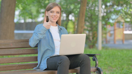 Thumbs Up by Young Woman Using Laptop while Sitting Outdoor on Benchの写真素材