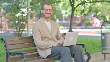 Middle Aged Man with Laptop Smiling at Camera while Sitting Outdoor on Benchの写真素材