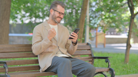 Middle Aged Man Celebrating Success on Phone while Sitting Outdoor on Benchの写真素材
