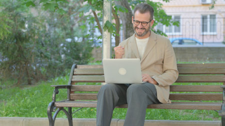 Young Man Celebrating Success on Laptop while Sitting Outdoorの写真素材