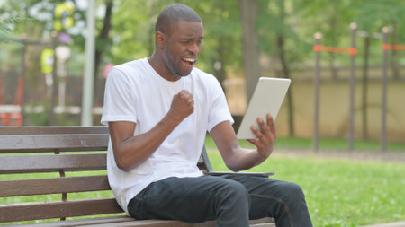 Outdoor Sitting Young African Man Celebrating Success on Tabletの写真素材