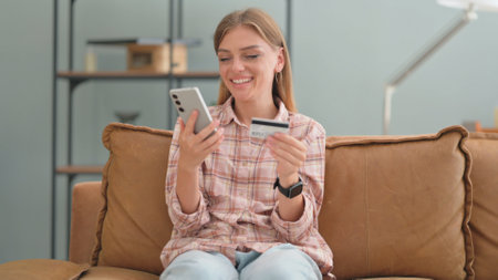 Young Woman Shopping Online with Phone while Sitting on Sofa at Homeの写真素材