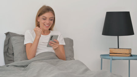 Young Woman Celebrating Success on Phone while Sitting in Bedの写真素材