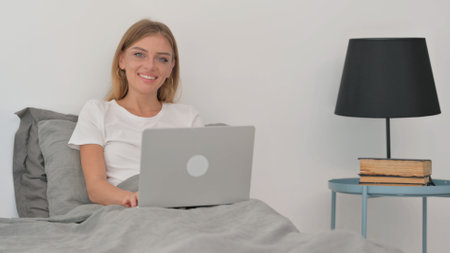 Smiling Young Woman Looking at Camera while in Bed with Laptopの写真素材