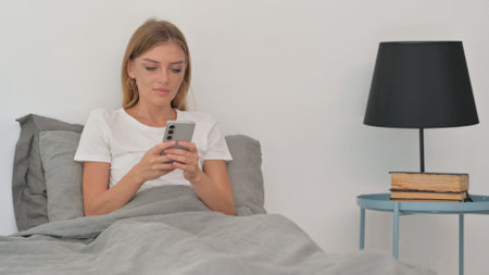 Young Woman Using Smartphone while Sitting in Bedの写真素材
