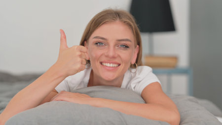 Close up of Young Woman doing Thumbs Up while Lying on Stomach in Bedの写真素材