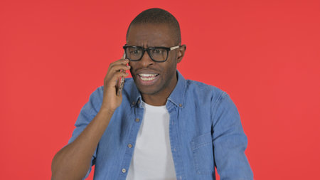 Angry Casual African Man Talking on Phone on Red Background, Portraitの写真素材