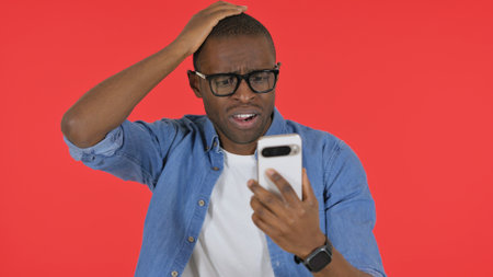Casual African Man Shocked by Loss on Phone on Red Background, Portraitの写真素材