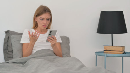 Young Woman Upset by Loss on Phone while Sitting in Bedの写真素材