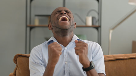 Portrait of Excited African Man Cheering Sitting on Sofa at Homeの写真素材