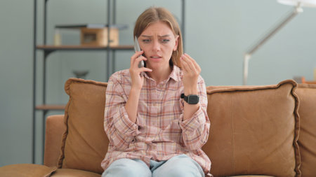Angry Young Woman Talking on Phone while Sitting on Sofa at Homeの写真素材