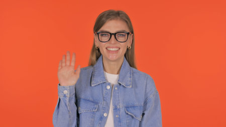 Casual Young Woman Waving Hand to Welcome on Orange Background, Portraitの写真素材
