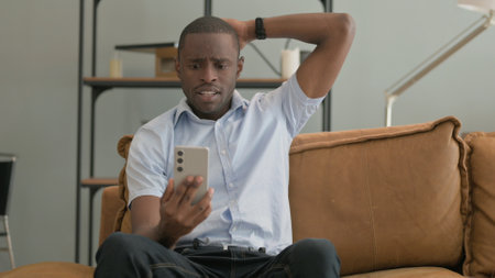 African Man Upset by Loss on Phone while Sitting in Bedの写真素材