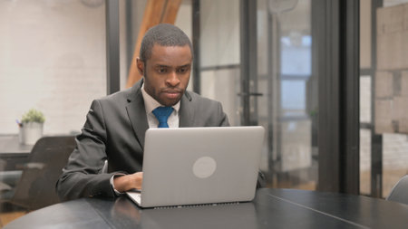 Serious African American businessman working on laptop in office. Serious black man in suit sitting...