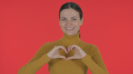Casual Young Woman Making Heart Sign with Hands on Red Backgroundの写真素材