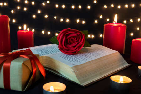 Romantic red rose on open book with gift box and candles in bokeh lights at night.