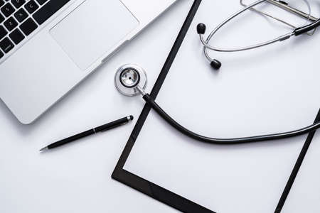 Top view of medical doctor table with stethoscope, clipboard, pen and laptop on white desk.