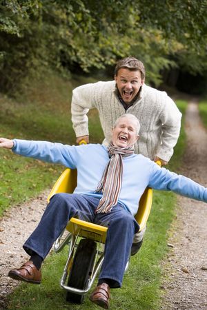 Man walking on path outdoors pushing other man in wheelbarrow and smiling (selective focus)の写真素材
