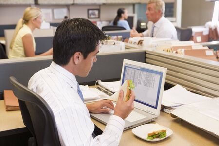 Businessman in cubicle at laptop eating sandwichの写真素材