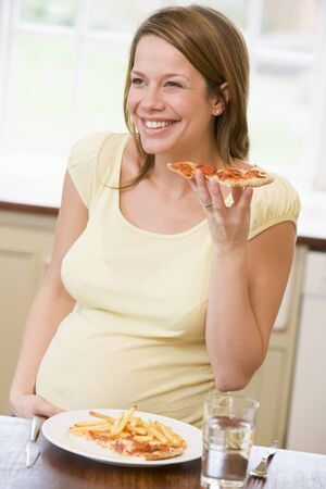 Pregnant woman in kitchen eating French fries and pizza smilingの写真素材