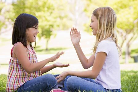 Two young girl friends sitting outdoors playing patty cake smilingの写真素材