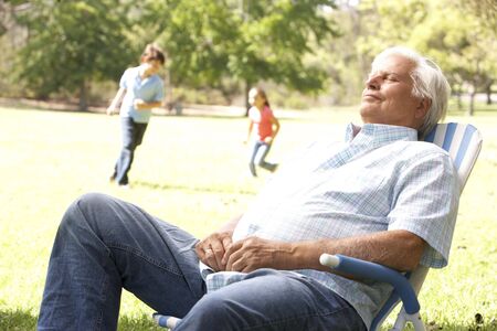 Senior Man Relaxing In Park With Grandchildren In Backgroundの写真素材