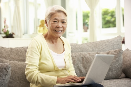 Senior Chinese Woman Using Laptop Whilst Relaxing On Sofa At Homeの写真素材