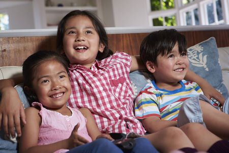 Three Asian Children Sitting On Sofa Watching TV Togetherの写真素材