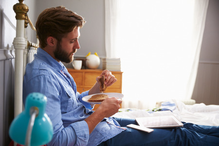 Man Eating Breakfast In Bed Whilst Using Mobile Phoneの写真素材