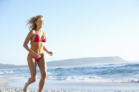 Young Woman Running Along Sandy Beach On Holiday Wearing Bikiniの写真素材