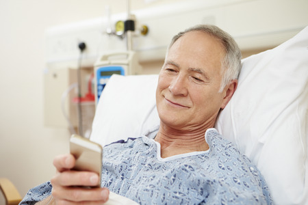 Senior Male Patient Using Mobile Phone In Hospital Bedの写真素材