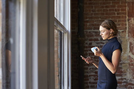 Businesswoman Checking Phone Standing By Office Windowの写真素材