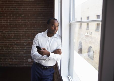 Businessman Checking Phone Standing By Office Windowの写真素材