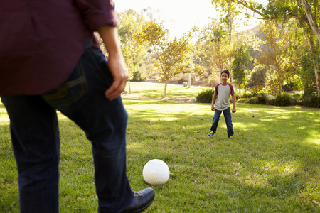 Dad kicking football to seven year old son in a park, cropの写真素材