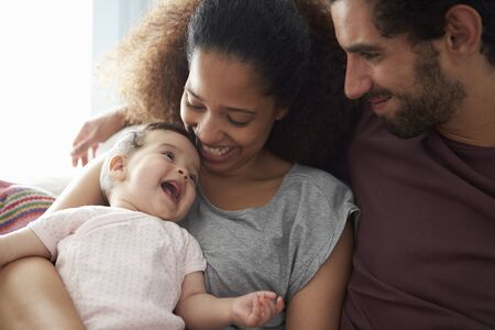 Parents Sitting On Sofa Cuddling Baby Daughter At Homeの写真素材