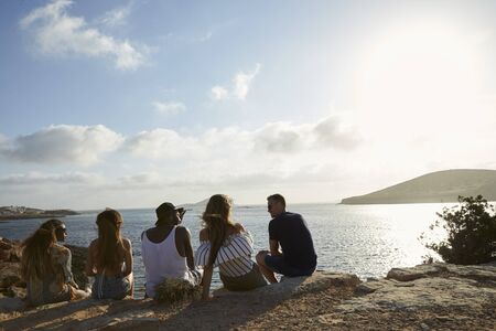 Rear View Of Friends Sitting On Cliff Watching Sunsetの写真素材
