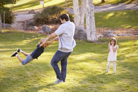 Father Swinging Son By His Arms In Park As Daughter Watchesの写真素材
