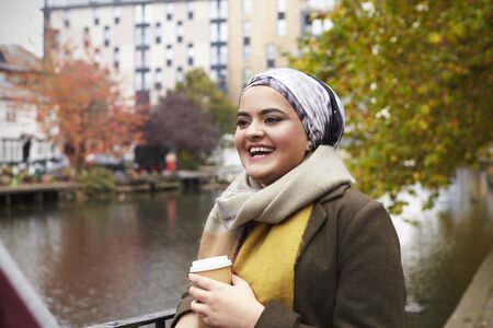 British Muslim Female With Takeaway Coffee By River In Cityの写真素材